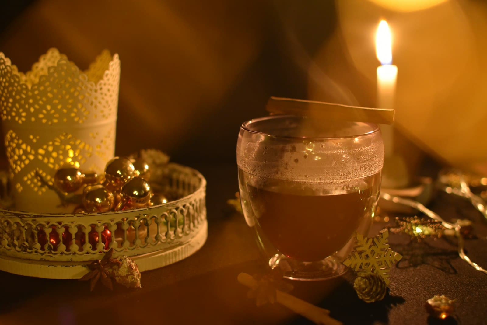 A warm spiced drink with a cinnamon stick beside a candle and gold ornaments, by candlelight
